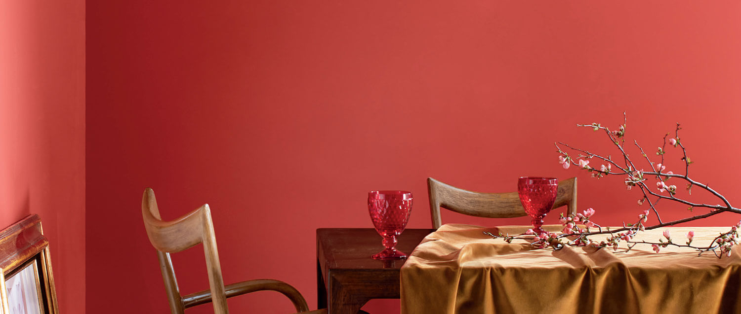 Dining room with coral tinged with pink painted wall, a table, chairs, cups and a branch with buds